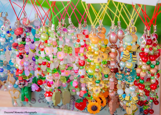 A close-up of many colorful beaded phone charms or keychains hanging from red and yellow strings against a blurred green background.