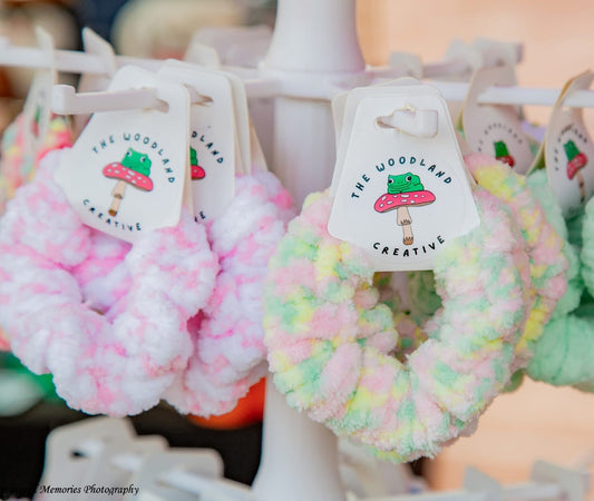 Three fluffy yarn scrunchies hanging from a white display stand, each with a paper tag reading 'The Woodland Creative.' The scrunchies are pastel pink, pastel yellow-green, and mint green.