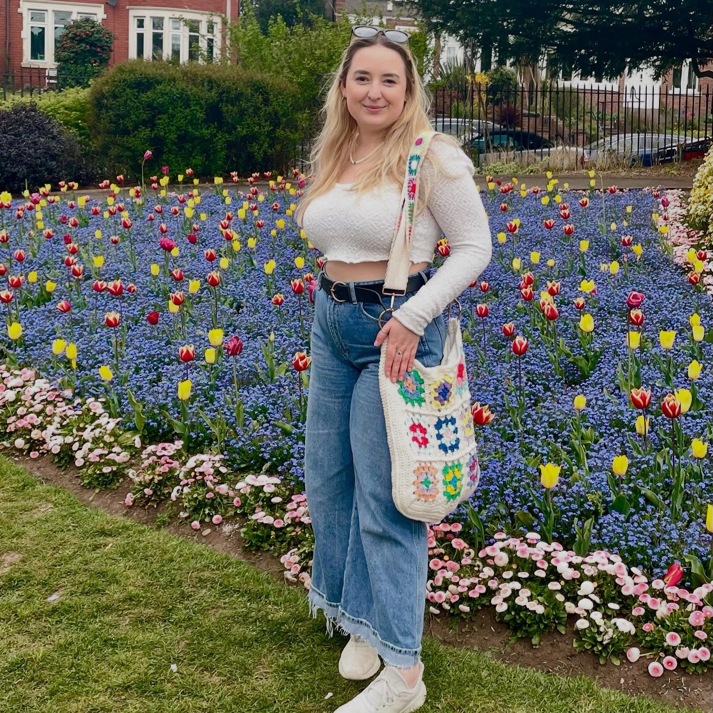 Woman standing in a garden with colorful flowers, wearing a white top, blue jeans, and a floral bag.