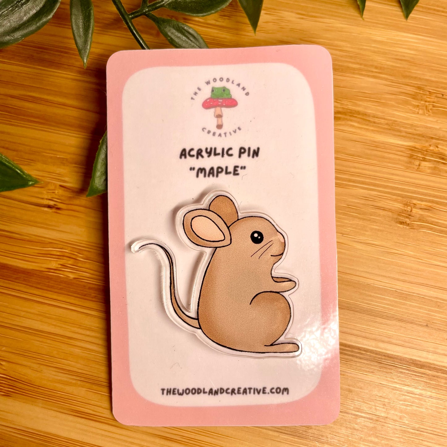 Acrylic pin of a mouse on a pink card with 'The Woodland Creative' branding, placed on a wooden surface.