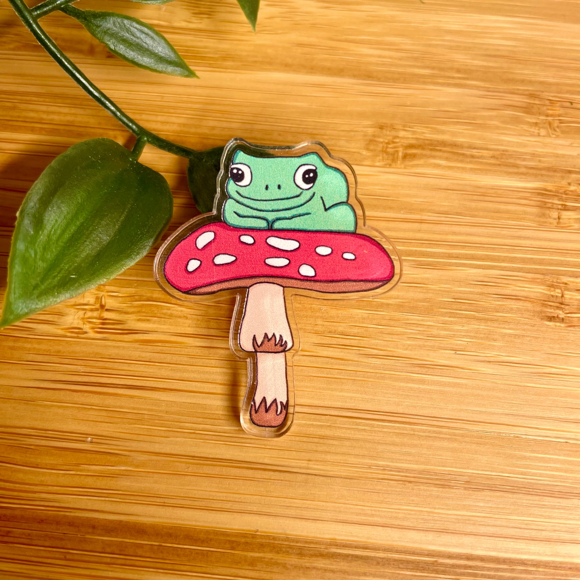 Frog on a mushroom pin badge on a wooden surface with leaves