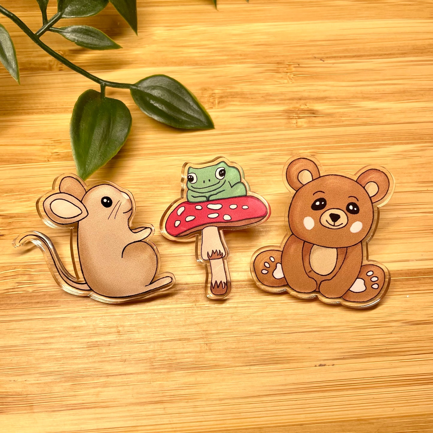 Three animal-themed pins (mouse, frog on a mushroom, bear) on a wooden surface with leaves.