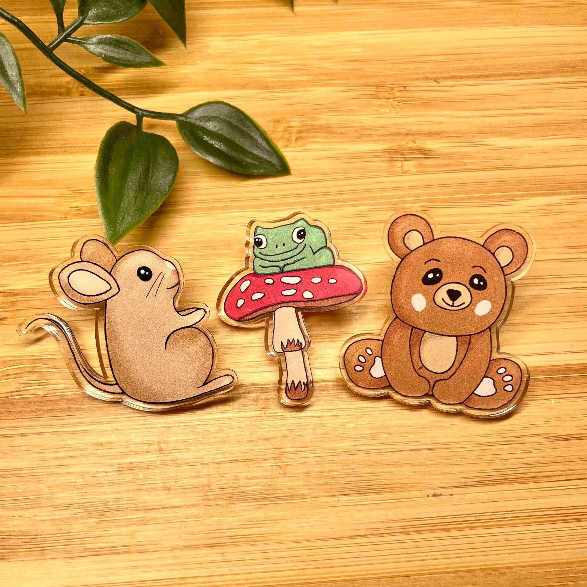 Three animal-themed pins (mouse, frog on a mushroom, bear) on a wooden surface with leaves.