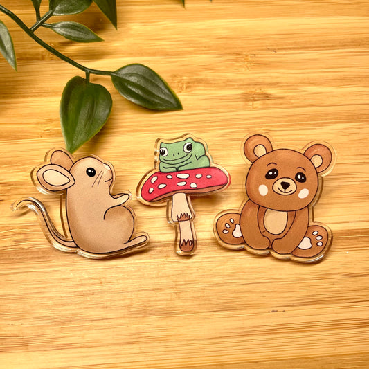 Three animal-themed pins (mouse, frog on a mushroom, bear) on a wooden surface with leaves.