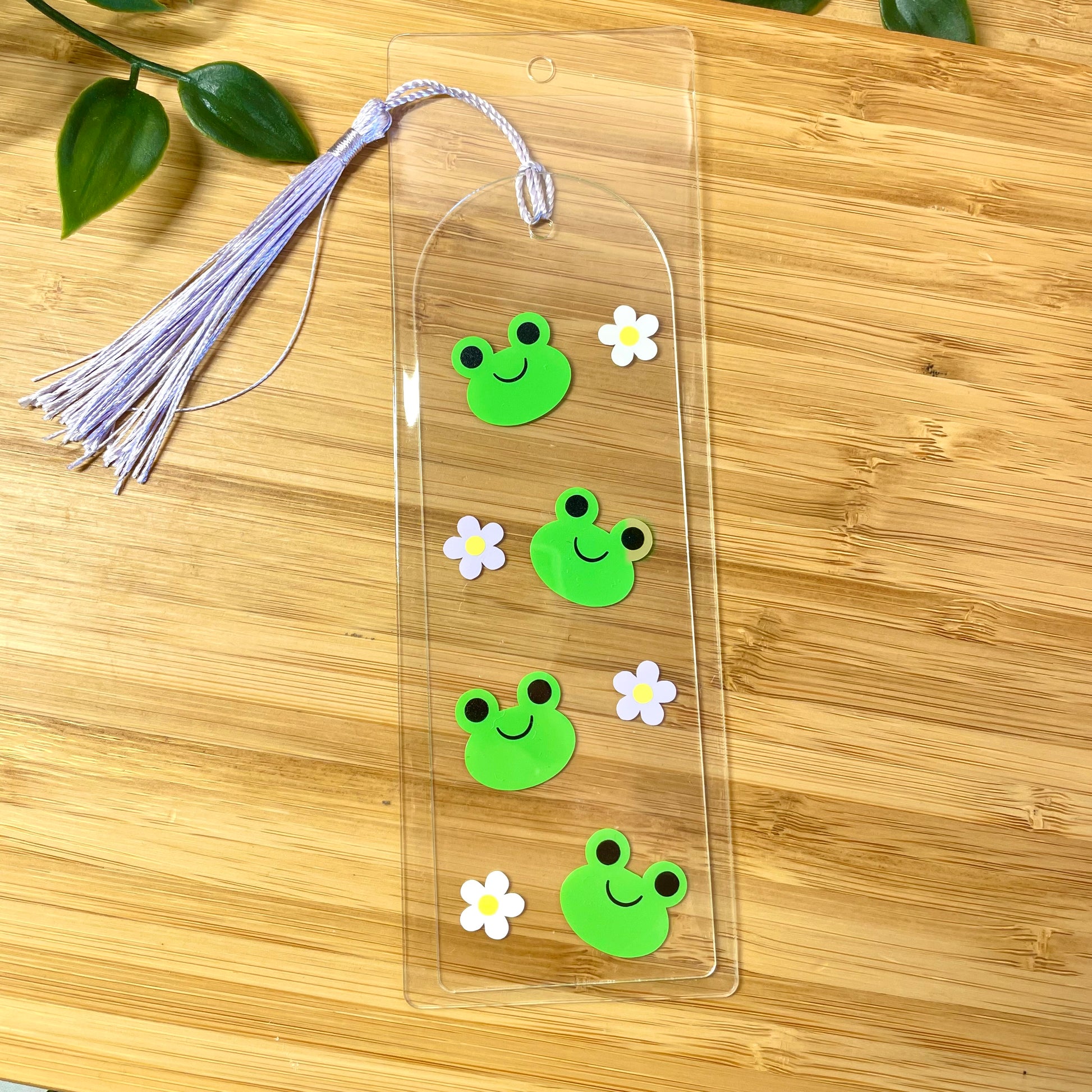 Clear bookmark with green frog stickers and white flowers on a wooden surface