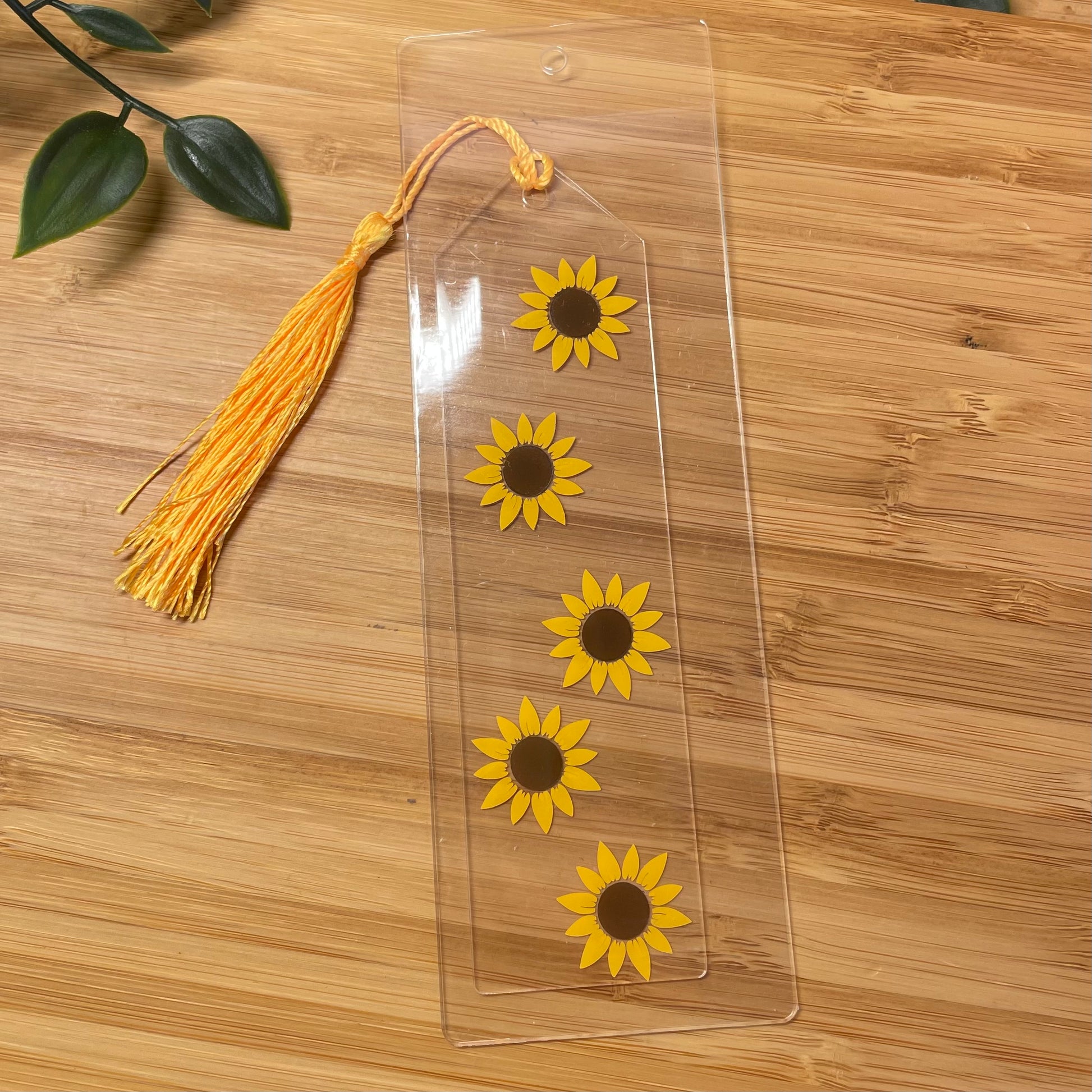 Clear bookmark with sunflower designs and a yellow tassel on a wooden surface.