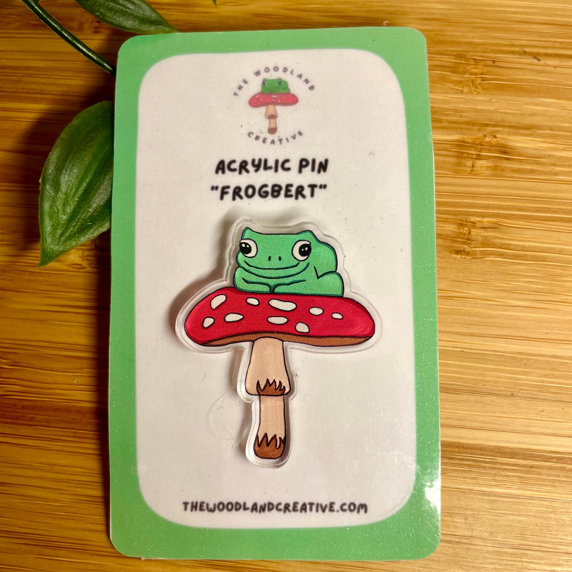 Acrylic pin of a frog on a mushroom with 'The Woodland Creative' branding on a wooden surface.