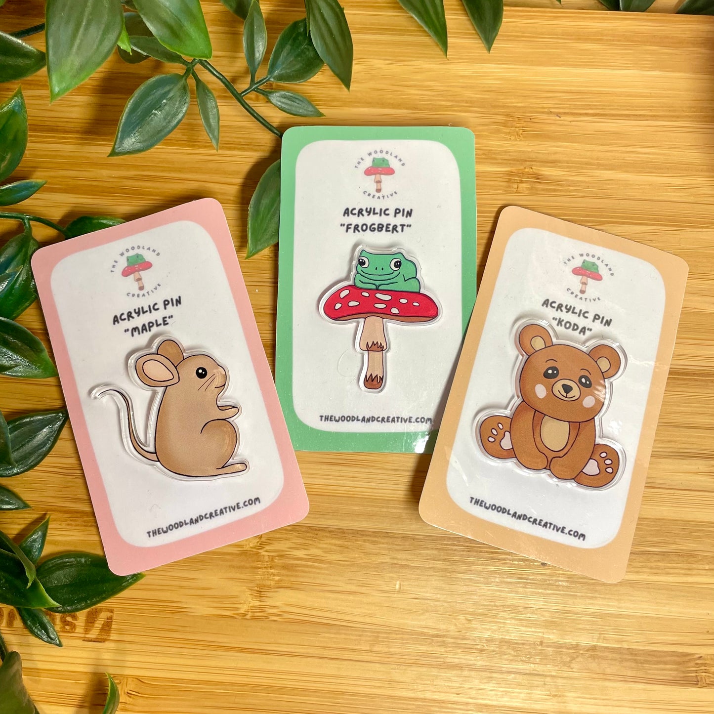 Three acrylic pins on cards with illustrations of a mouse, frog, and bear on a wooden surface.